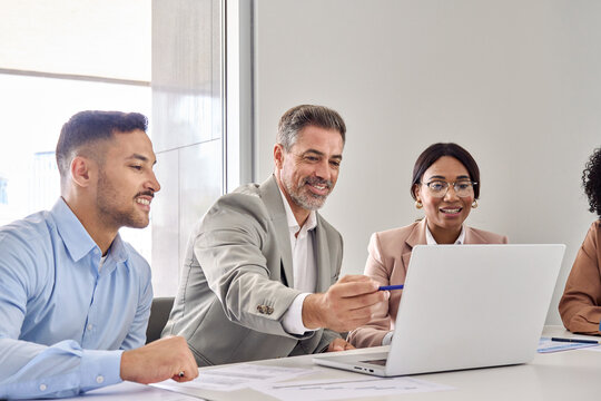 Happy older executive manager teaching busy business people workers board diverse team showing online project work at laptop using computer having discussion at office professional group meeting.