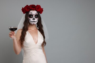 Young woman in scary bride costume with sugar skull makeup, flower crown and glass of wine on light grey background, space for text. Halloween celebration