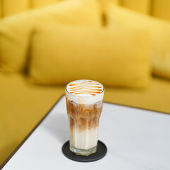 Iced Caramel Machiatto. Soft focus on the caramel on top of cream foam, blurred background of yellow couch and pillows