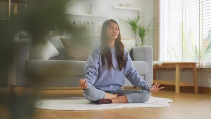 Happy young Asian woman practicing yoga and meditation at home sitting on floor in living room in lotus position and relaxing with closed eyes. Mindful meditation and wellbeing concept - Powered by Adobe