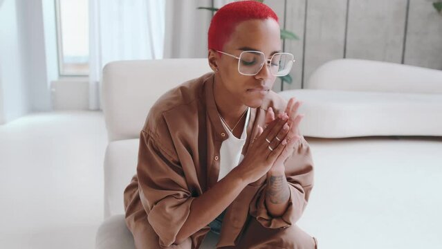 Young anxious African American woman is nervous and rubbing palms resting chin in hands waiting for something important dressed in casual clothes sits in living room with white stylish sofas.