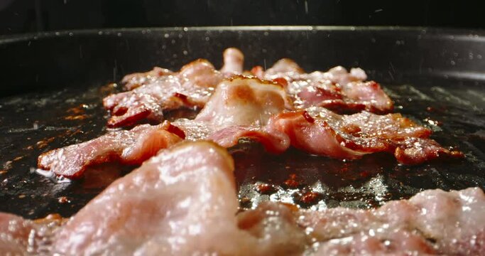 Frying pork bacon in pan side view close up slow motion. Splashes of sizzling oil. Pan-fried bacon slices capture the essence of sizzling goodness, ready to be devoured with delight. Fried to perfect