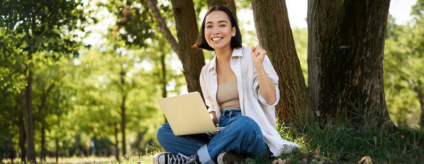 Enthusiastic young asian girl, sitting with laptop beside tree in green sunny park, celebrating, triumphing and smiling