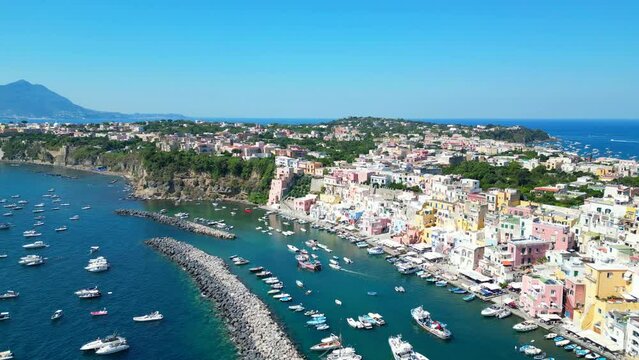 Splendid aerial pullback shot from Panoramica sulla Corricella on Procida Island (Isola di Procida) with breakwaters, boats on the Tyrrhenian Sea, pastel buildings, greenery, Naples' gulf, Vesuvius