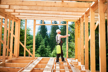 Laborer constructing wooden frame house near forest. Man treating woods, applying fire retardant using sprayer, while dressed in protective suit, helmet. Concept of modern eco-friendly construction. - obrazy, fototapety, plakaty