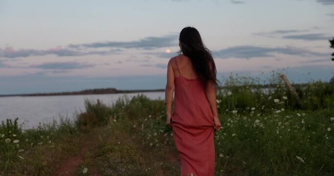 Happy young woman wearing dress outdoors in field overlooking ocean at sunset - hippy vibes