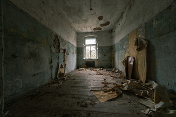 Fototapeta na wymiar Interior of abandoned room with peeling painted walls damaged wooden furniture garbage on dirty floor with crumbling ceiling showing iron rods with glass window in daylight