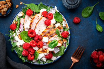 Tasty salad with raspberries, grilled chicken, cheese, onion, walnuts, spinach and arugula, blue table background, top view