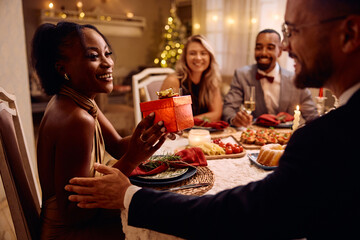 Happy black woman receives Christmas present during dinner party.