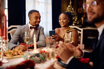 Happy black couple using cell phone during Christmas dinner party.