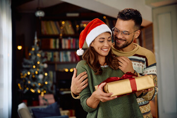 Happy man surprising his girlfriend with Christmas present at home. - obrazy, fototapety, plakaty