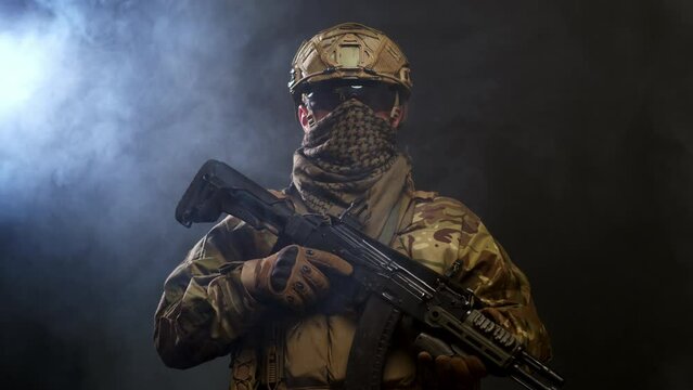 A courageous soldier in a protective combat uniform with a machine gun standing on a dark background, shrouded in smoke. War and military concept.