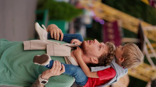 Vertical video of a father in a Green T-shirt and his little boy, his blond son with blue eyes in a red T-shirt sits on his shoulders and does not look at the rides in an amusement park during the day