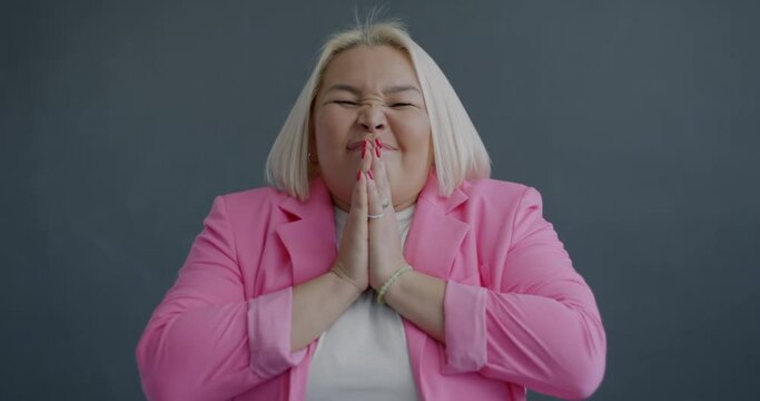 Slow motion portrait of desperate young Asian woman begging making prayer gesture looking at camera on gray background. People and emotion concept.