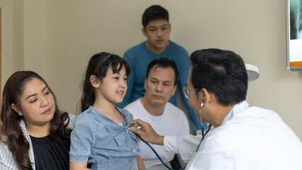 Fototapeta premium Doctor uses stethoscope to check a girl's lung in his clinic. Doctor discusses with a girl's family for a medical treatment program.