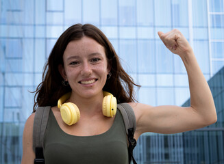 Young redhead woman with freckles showing biceps looking at camera in a city. Empowered young...