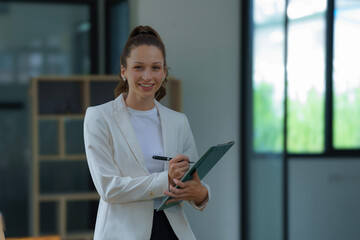A businesswoman stands holding a document file and looking at a camera in an office.