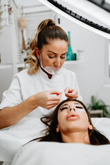 Beautiful young adult woman receiving professional eyelash makeup treatment. Cosmetician is smiling and looking at camera. Beauty and cosmetics business.