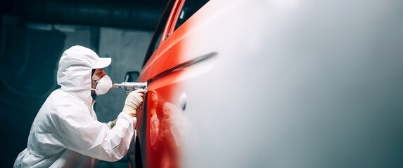 A car painter in a white protective suit paints a car with red spray paint. Copy space. Banner. - obrazy, fototapety, plakaty