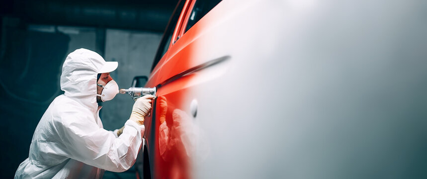 A car painter in a white protective suit paints a car with red spray paint. Copy space. Banner. - Powered by Adobe