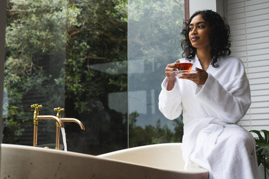 Happy biracial woman in bathrobe sitting on bath with tea and looking out window in sunny bathroom