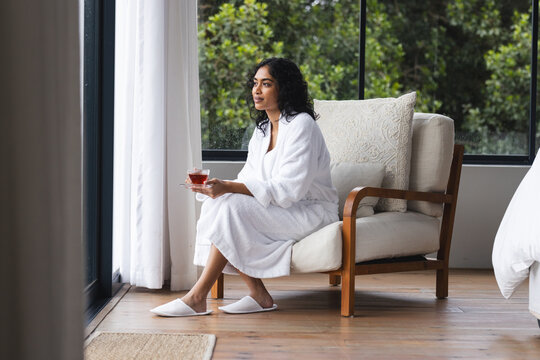 Pensive biracial woman in bathrobe sitting with cup of tea and looking ahead in sunny room at home