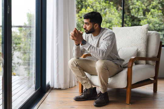Pensive biracial man sitting on armchair and looking out window in sunny living room