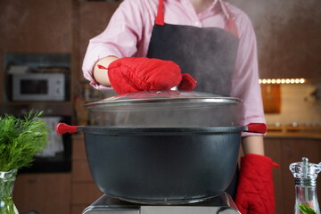 Woman  in oven heat gloves open lid of black pot with boiling water . Steam over pot