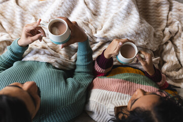 Biracial couple sitting on sofa under blanket and drinking cocoa in living room at home