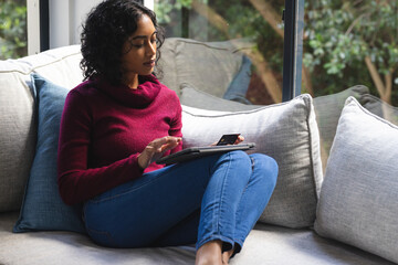 Happy biracial man sitting on sofa, using credit card and tablet in sunny living room