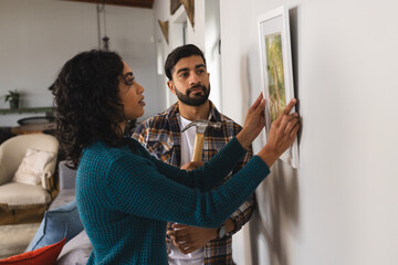 Focused biracial couple hanging picture on wall in living room at home