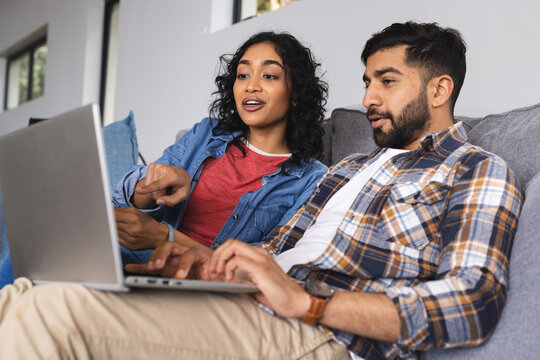 Happy biracial couple sitting on sofa and using laptop in living room at home