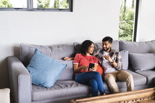 Happy biracial couple sitting on sofa and using smartphone in living room at home