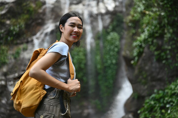 Beautiful young woman tourist with backpack standing in front of the tropical waterfall