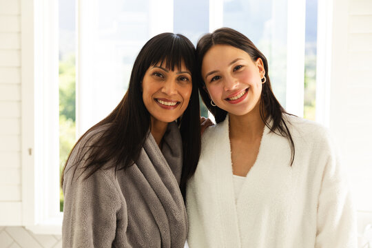 Happy diverse mother and daughter wearing bathrobes and embracing in bathroom at home, copy space