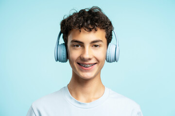 Portrait of attractive smiling boy with dental braces wearing wireless headphones isolated on blue...
