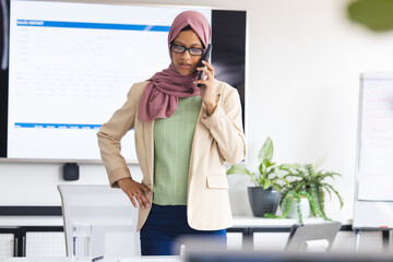Busy biracial casual businesswoman with hijab talking on smartphone in meeting office