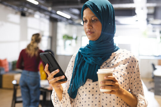 Busy biracial casual businesswoman with hijab using smartphone over colleagues working in office