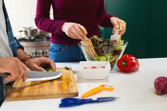 Midsection of caucasian couple chopping vegetables and tossing salad leaves in kitchen