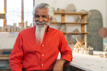 Happy biracial senior man with long beard sitting and smiling in pottery studio