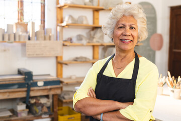 Happy biracial senior female potter with gray hair, standing and smiling in pottery studio