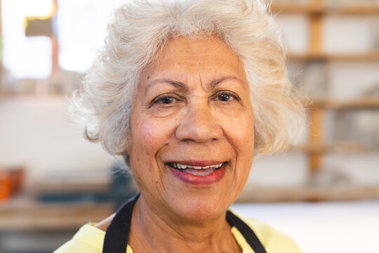 Happy biracial senior female potter with gray hair, looking on camera and smiling in pottery studio