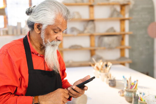 Focused biracial senior potter with long beard using smartphone in pottery studio