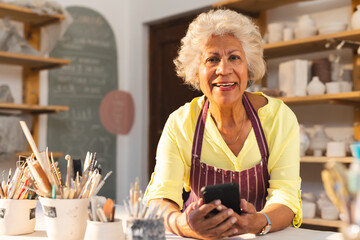 Happy biracial senior female potter with gray hair, using smartphone and smiling in pottery studio