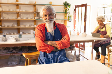 Focused biracial senior potter with long beard sitting and smiling in pottery studio