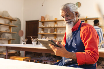 Focused biracial senior potter with long beard using tablet in pottery studio
