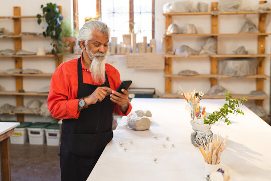 Focused biracial senior potter with long beard using smartphone in pottery studio