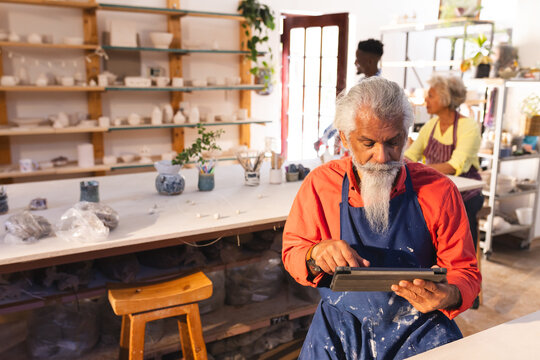 Focused biracial senior potter with long beard using tablet in pottery studio