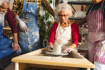 Happy biracial senior female potter with others, using potter's wheel in pottery studio