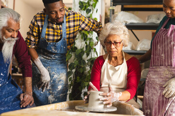 Focused biracial senior female potter with others, using potter's wheel in pottery studio